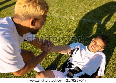 High angle view of caucasian player assisting injured teammate lying on field in getting up. Match, playground, summer, pain, unaltered, soccer, sport, teamwork, togetherness and competition.