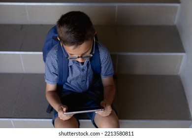 High Angle View Of A Caucasian Schoolboy Wearing Glasses, Shorts And A Rucksack Sitting On Steps At Elementary School Using A Tablet Computer