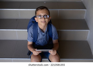 High Angle View Of A Caucasian Schoolboy Wearing Glasses, Shorts And A Rucksack Sitting On Steps At Elementary School Using A Tablet Computer, Looking Up To Camera And Smiling