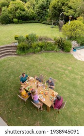 High Angle View Of Caucasian Three Generation Family Sitting At Table Eating Meal In Garden. Three Generation Family Celebrating Eating Outdoors Together.