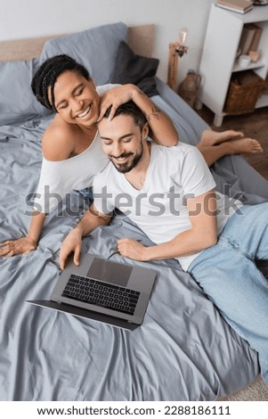 high angle view of cheerful interracial couple in white t-shirts watching film on laptop on bed at home
