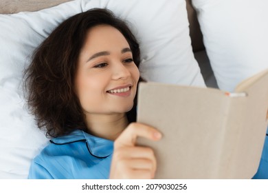 High Angle View Of Cheerful Young  Woman Reading Book In Bed