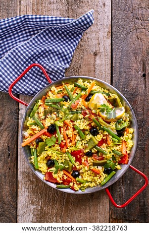High Angle View of Colorful and Fresh Vegetarian Paella Spanish Rice Dish Served in Pan with Red Handles and with Blue and White Checkered Linen Napkin on Rustic Wooden Table