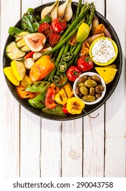 High Angle View Of Colorful Grilled Vegetable Bounty On Cast Iron Pan With Olives And Dip Resting On White Painted Wooden Picnic Table With Copy Space