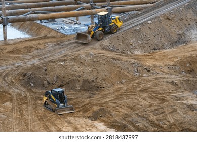 High Angle View of Construction Machinery Working at Large Earth Excavation Site Featuring Yellow Front Loader and Skid Steer Loader Clearing Dirt and Moving Soil for Foundation Development