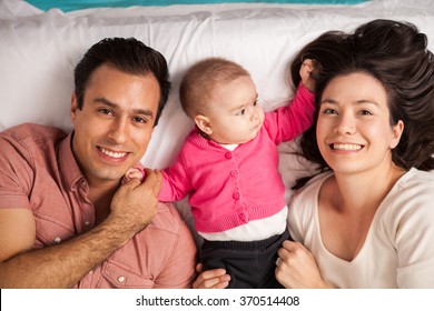 High Angle View Of A Couple Of Attractive Young Hispanic Parents Lying In A Bed With Their Cute Baby Girl