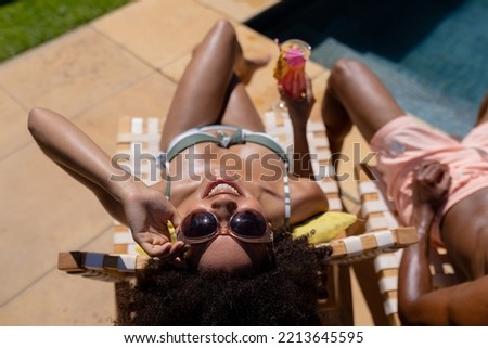 High angle view of a  couple sitting in the garden by a swimming pool sunbathing on sunloungers wearing beachwear, the woman in sunglasses, holding a cocktail and looking to camera smiling