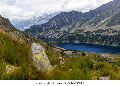 High angle view of a deep blue alpine lake surrounded by rocky mountain peaks and green slopes under cloudy summer sky.