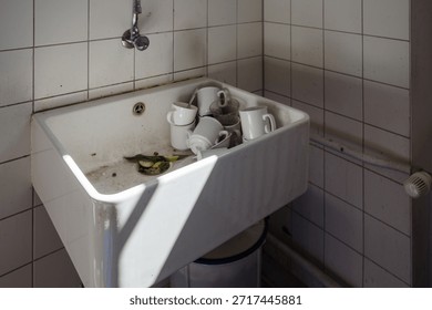 A high angle view of a deep white ceramic utility sink located in a tiled corner. The basin is cluttered with dirty white coffee mugs, drinking glasses, and food waste including cucumber slices and cigarette ash