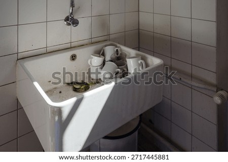 A high angle view of a deep white ceramic utility sink located in a tiled corner. The basin is cluttered with dirty white coffee mugs, drinking glasses, and food waste including cucumber slices and cigarette ash