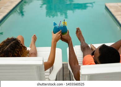 High Angle View Of Diverse Couple Toasting Glasses Of Cocktail While Relaxing On A Sun Lounger Near Swimming Pool At The Backyard Of Home. Summer Fun At Home By The Swimming Pool