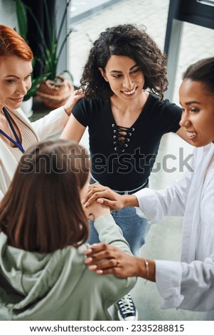 high angle view of diverse group of pleased multicultural women joining hands near positive motivation coach during session in consulting room, moral support and mental wellness concept