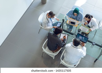 High angle view of diverse medical team discussing with each other at table in hospital. Coffee cup, medical folders, clipboard, digital tablet and laptop are on the table. - Powered by Shutterstock