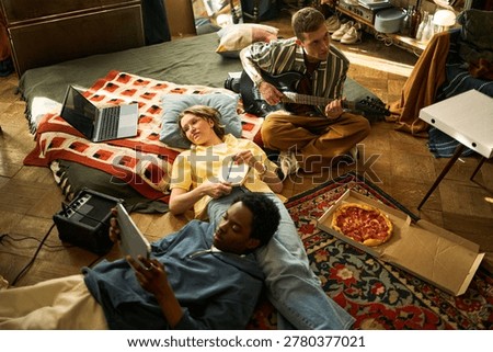 High angle view of diverse young adults collaborating on music project, playing guitar, using laptop and tablet, taking notes. Ideal for creative teamwork, youth lifestyle marketing