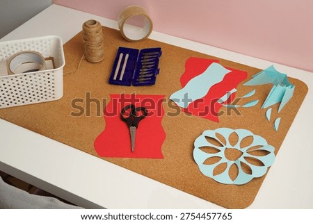 High angle view of a DIY crafting project. A cork board holds red and blue paper cutouts, scissors, and knives. This creative workspace evokes a feeling of hobby and handmade artistry. (188 chars)
