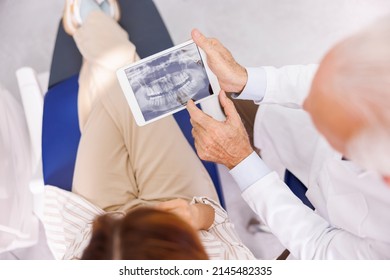 High Angle View Of Doctor Showing Jaw X-ray On Tablet Computer To Patient Sitting At Dental Chair At Dentist Office