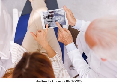 High Angle View Of Doctor Showing Jaw X-ray On Tablet Computer To Patient Sitting At Dental Chair At Dentist Office