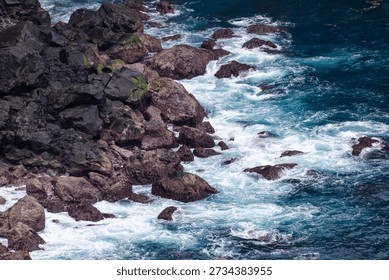 High angle view of dramatic seascape featuring foaming blue water turbulence surrounding rough, moss-covered coastal boulders.