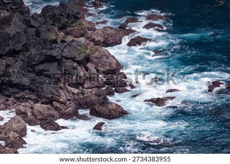 High angle view of dramatic seascape featuring foaming blue water turbulence surrounding rough, moss-covered coastal boulders.