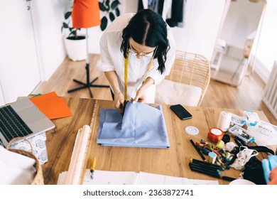 High angle view of female dressmaker standing at wooden table with laptop smartphone sewing material colorful pencils while cutting fabric with scissors in creative workshop - Powered by Shutterstock