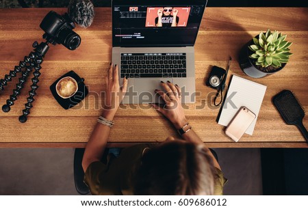 High angle view of female vlogger editing video on laptop. Young woman working on computer with cameras and accessories on table.