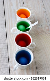 High angle view of four cups with dye for Easter eggs. The cups are filled with of yellow, blue, red and green dye. There are eggs in each cup on a rustic farmhouse style kitchen table.