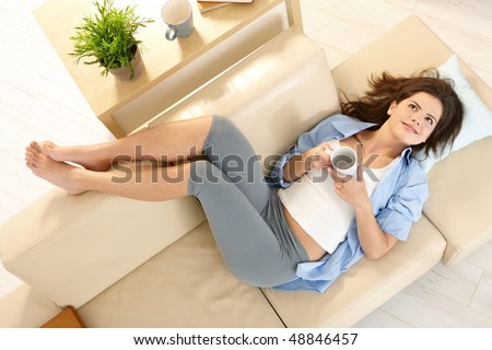 In high angle view girl lying on living room couch with feet up, smiling, holding mug in two hands.
