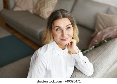 High angle view of gorgeous mature 60 year old woman with beautiful wise eyes and joyful smile relaxing on comfortable grey couch wearing white shirt, staring at camera with happy positive look