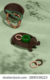 A high angle view of green jade rings and beaded bracelets displayed on carved wooden holders against a soft sage green background with natural shadows. Jewelry pieces are arranged on a green marble-style surface with decorative hedgehog animal case figurine and natural accessories nearby. Rings, beads and sculptural objects placed in a balanced still life composition. Dark green, brown and muted stone tones create a rich earthy palette. Glossy jewelry surfaces, rough decorative textures and soft shadows visible in the frame. Decorative tabletop photograph with calm styling and curated arrangement. Still life centered on accessories, figurine, natural decor and home display details. Minimal composition with artistic balance and soft spa-inspired colors.
