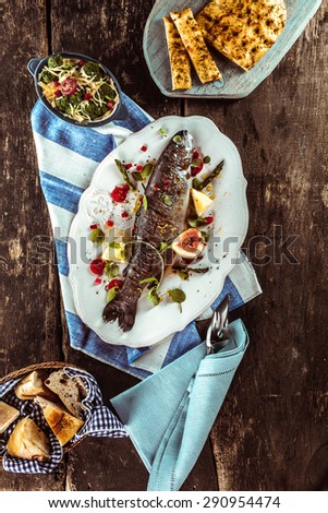 High Angle View of Grilled and Garnished Whole Fish on Wooden Table Surrounded by Other Dishes and Linen Napkins