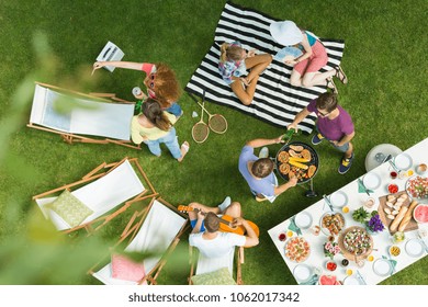 High Angle View Of Group Of People Having Backyard Barbecue Party With Grill, Guitar, Deck Chairs And Delicious Food On The Table