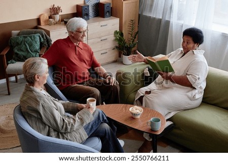 High angle view at group of senior people enjoying book club discussion in cozy living room at retirement community with African American woman reading aloud