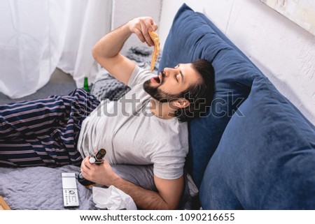 high angle view of handsome loner eating pizza and holding bottle of beer in bedroom