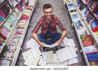 High angle view of handsome student reading a book, looking at camera and smiling while sitting on the floor among open books in the bookshop - Shutterstock ID 2221121407