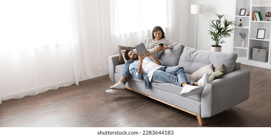 High angle view of happy smiling multiracial millennial couple relaxing together on couch at home, handsome arab guy wearing glasses using digital tablet lying on indian lady laps, copy space, banner - Powered by Shutterstock