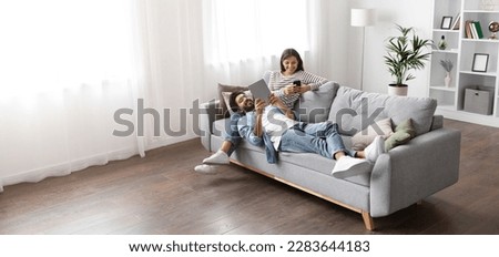 High angle view of happy smiling multiracial millennial couple relaxing together on couch at home, handsome arab guy wearing glasses using digital tablet lying on indian lady laps, copy space, banner