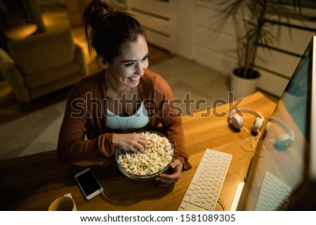 High angle view of happy woman relaxing in the evening and eating popcorn while watching movie on her computer at home. 