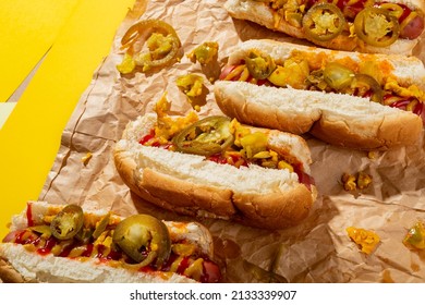 High Angle View Of Hot Dogs With Sauces And Jalapeno On Brown Wax Paper Over Yellow Background. Unaltered, Unhealthy Food, Meat, Bread, Sausage, Sauce, Jalapeno And Fast Food.