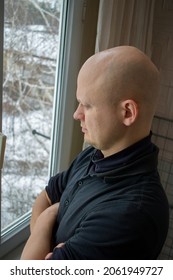 High angle view image of bald man dressed in black sweatshirt standing near window and looking on the street in winter. Serious and thoughtful bald man standing near window in winter at his apartment.