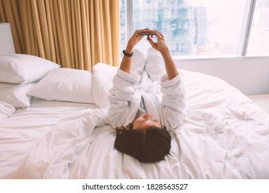 High angle view image of a woman wearing white bathrobe lying down in bed facing to bedroom ceiling and using mobile phone with city view through window.