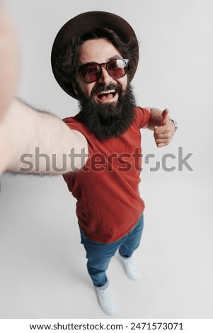 High angle view of a joyful bearded man wearing a hat and sunglasses, giving a thumbs up in a friendly gesture. His cheerful demeanour and trendy casual attire convey a sense of happiness, positivity.
