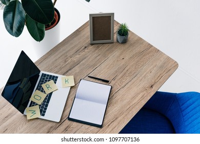 High Angle View Of Laptop With Blank Screen, Lettering Work On Post Its, Empty Textbook, Photo Frame And Potted Plant On Table