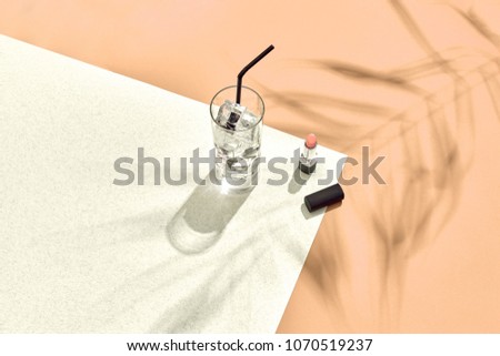 High angle view of lipstick and drinking glass with ice on table. Beige and white background with shadow from a palm leaf