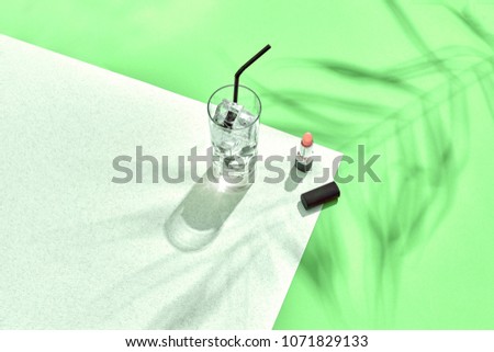 High angle view of lipstick and drinking glass with ice on table. Green and white background with shadow from a palm leaf