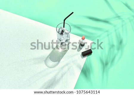 High angle view of lipstick and drinking glass with ice on table. Blue and white background with shadow from a palm leaf
