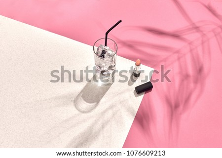 High angle view of lipstick and drinking glass with ice on table. Pink and white background with shadow from a palm leaf