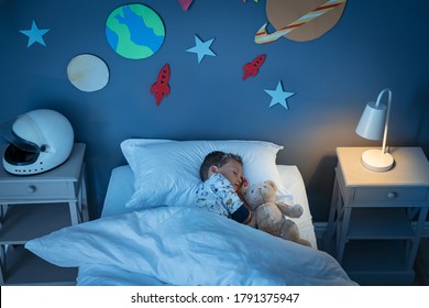 High angle view of little boy dreaming of becoming an astronaut while sleeping with teddy bear in space decorated room. Top view of dreamer child sleeping on bed during the night and the light on.