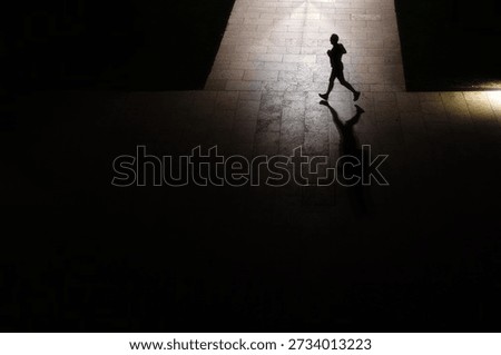 High angle view of a lone runner silhouette on a lit pavement in a dark park. Urban night fitness and healthy lifestyle.