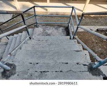 High angle view looking down weathered outdoor concrete stairs with a rusty metal handrail. The staircase has a landing, turning downwards alongside a stone wall with scattered dry leaves on the steps.