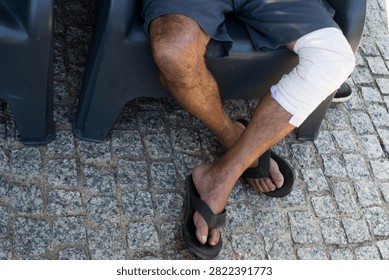 High angle view looking down at a man's crossed legs sitting outdoors, featuring one leg wrapped with a white compression bandage and flip-flops on cobblestone.
